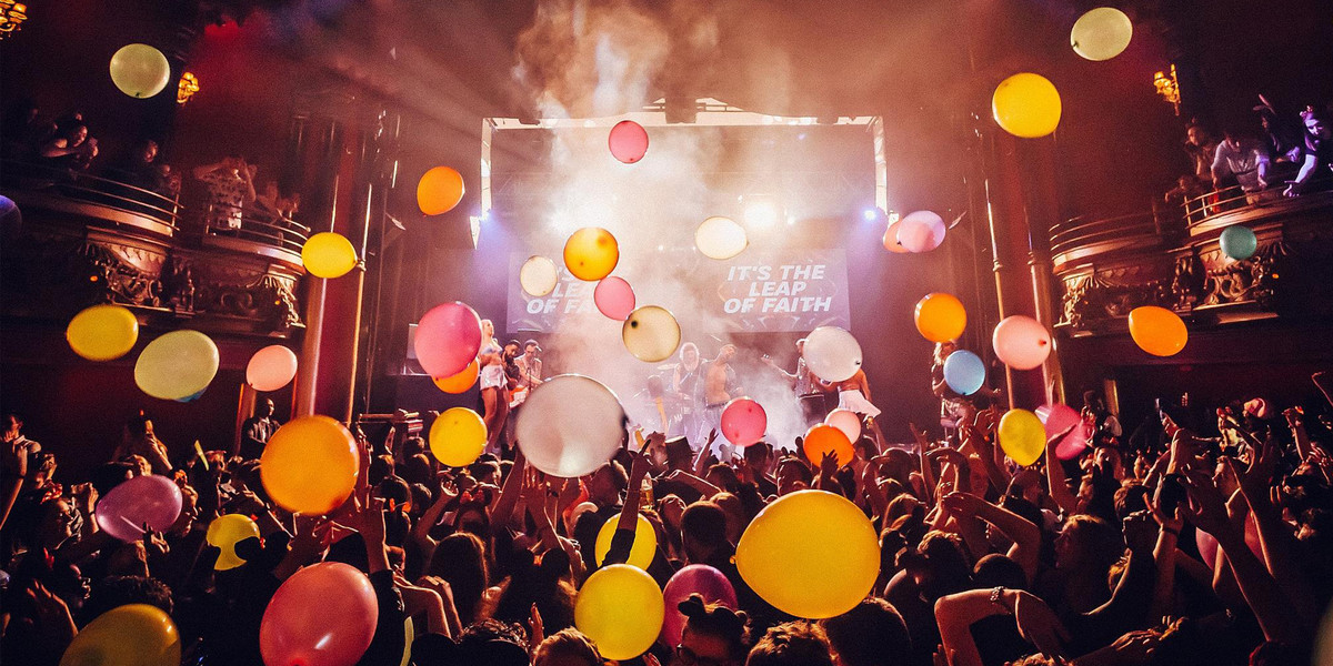 A crowd of tightly packed people crowd the lower foreground, hands aloft. There are balloons falling from the ceiling, some of the members of the crowd appear to be bouncing them upwards. Above the crowd to each side are ornate royal boxes with a couple of people standing in them. The centre is filled with a well-lit stage, slightly obscured by the falling balloons and some haze diffusing some warm white stage lights. On the left of the stage is a femme-presenting person in white shorts and a pale cropped top, next to them are two masc-presenting people, one with dark short hair and a dark beard, the other is clean shaven with lighter hair playing a red and white guitar. Centre stage we can see a masc-presenting person wearing jeans and an open-shirt showing a bare chest, head tilted to the left in the appearance of ecstasy, behind him we see a masc-appearing drummer with fair shoulder length hair wearing a dark headband and black tank. To the right we see a masc-appearing person wearing a pale shirt playing a bass guitar, standing next to someone wearing a short, full white skirt that appears to be swaying in motion, both are half-obscured by a balloon in the foreground. Above the musicians appear two tv screens with identical text on them that reads "It's the leap of faith".