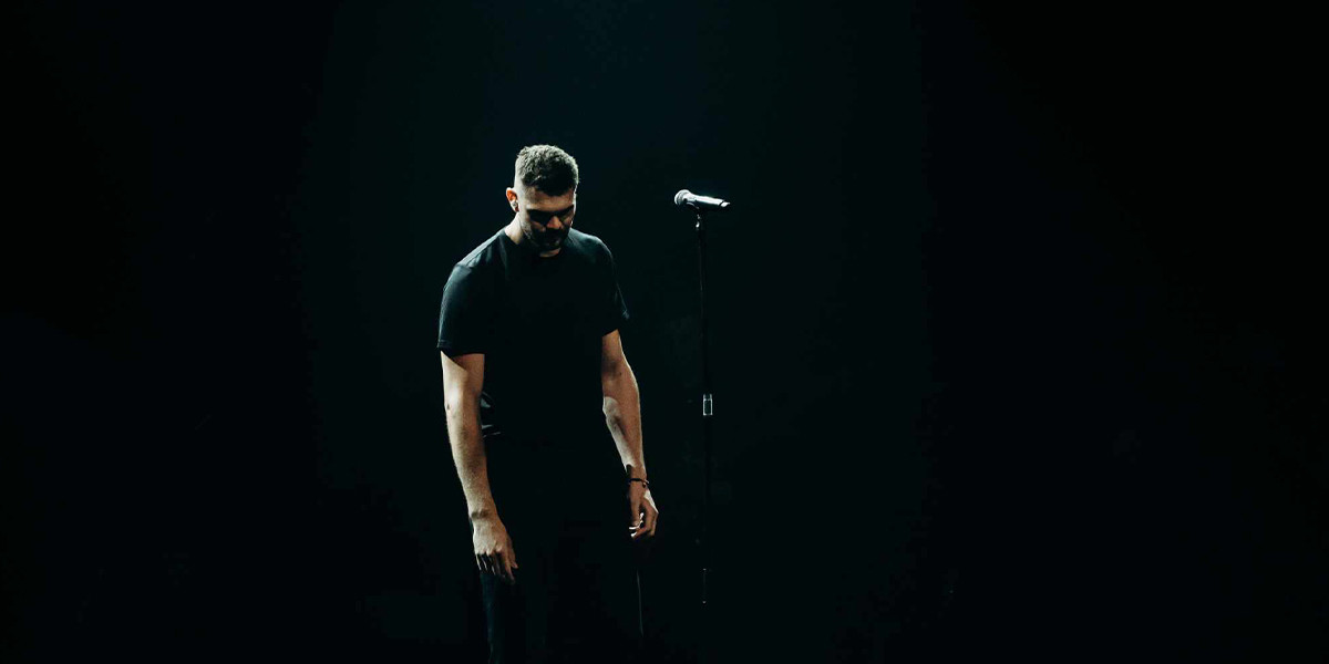A tall man in  black T-shirt stands on a dimly lits stage with his head pointed down.