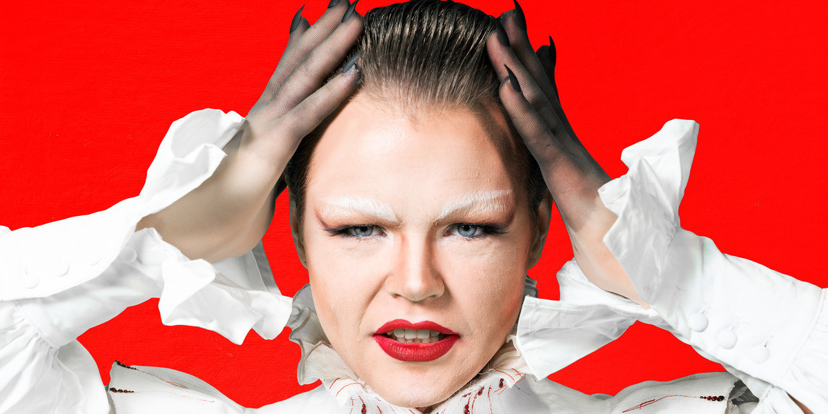 Florian Wild, a caucasian man with blue eyes and brown hair, is looking directly into the camera, with his hands on his head. He has drag queen makeup on, with white eyebrows and a red lip. He is dressed in a white blouse and a tall white vampire collar with red diamonte accents like blood droplets. He has black nails on his hands. The background is red.