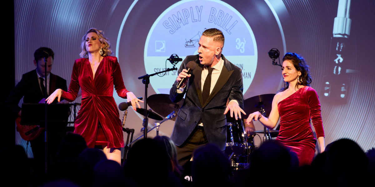 A taller blonde woman, and shorter brunette woman stand on either side of a brunette man who is singing. The two women are wearing short maroon velvet dresses, and are dancing the twist. The man is dancing also. Behind them, a guitarist plays and there is a drumkit. There is an audiovisual screen featuring a record spinning, that says Simply Brill.
