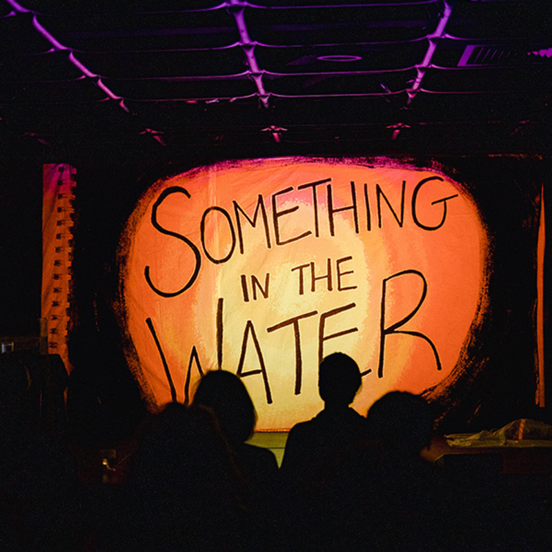 A projection screen in a theatre reads a hand-drawn title: "Something in the Water"