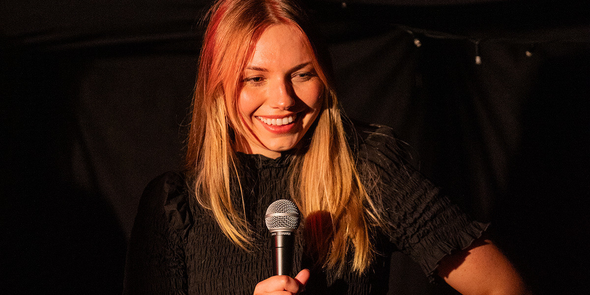 Comedian Jodie Sloan smiling at crowd while performing standup comedy into a mic.