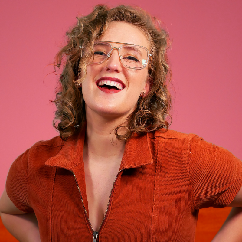 Millicent Sarre is Opinionated - Mim, a white woman with curly, dark blonde hair stands with her hands on hips, smiling triumphantly with her mouth open. She is wearing a burnt-orange corduroy jumpsuit and oversized glasses, and stands against a muted pink background.