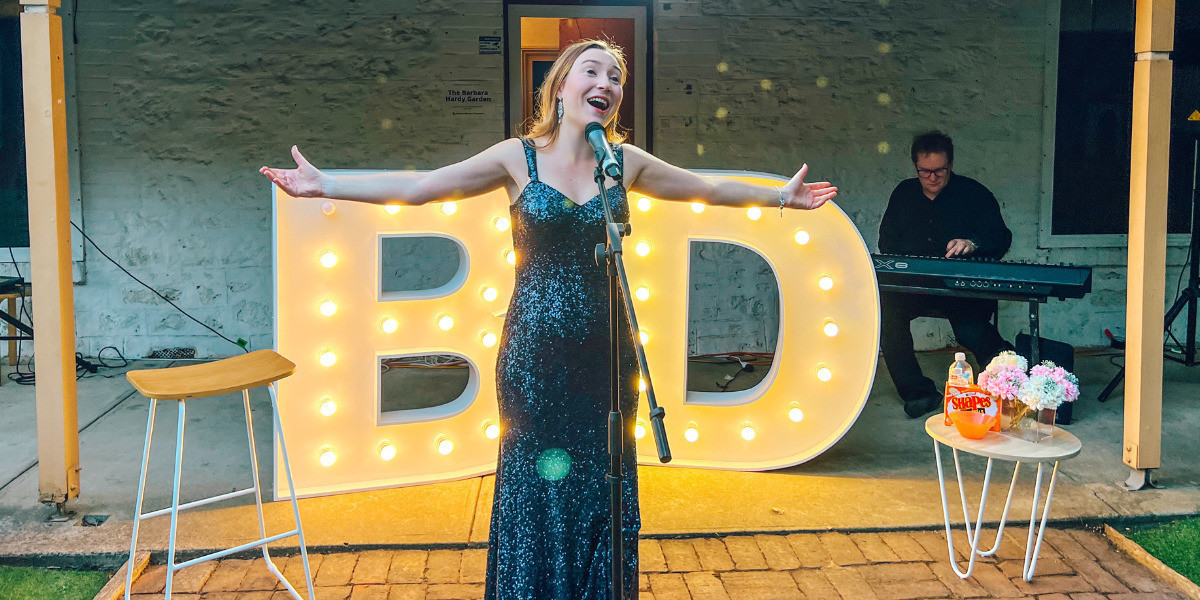 A performer sings into a microphone, in front of a light up letter B and D. She holds her arms out wide.