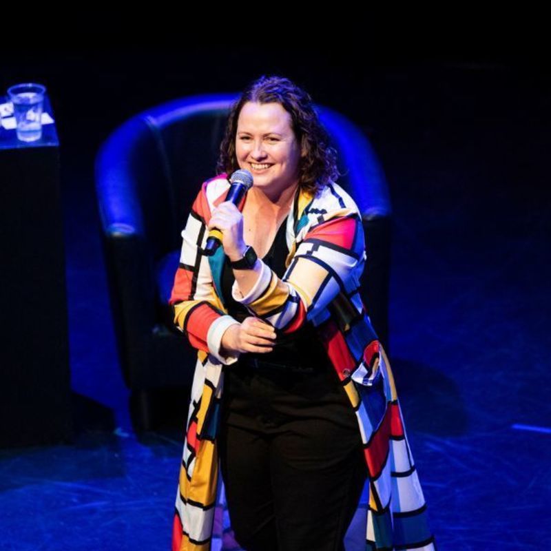 KC performing on stage, wearing black pants and a top with a colourful long jacket in Piet Mondrian’s famous geometric print. She holds a microphone and is smiling. In the background beyond the spotlight, the stage is washed with blue light. There is a comfortable chair, and a side table piled with books and a bottle and glass of water.