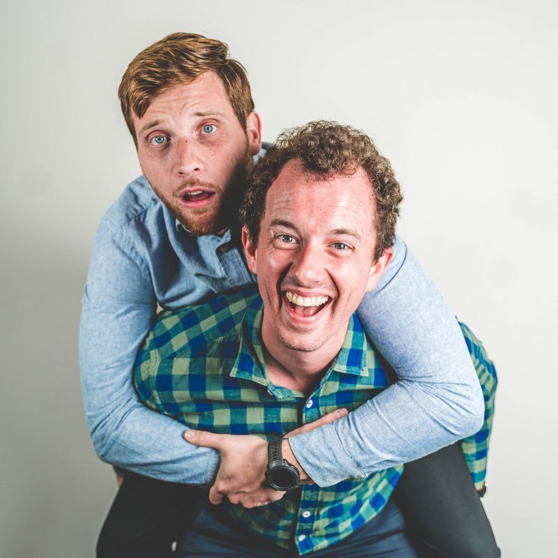 Workmates - Image of David Arcidiaco and Robbie Greenwell on a white background. Robbie is giving David a piggyback ride and looking very happy, while David looks very apprehensive.