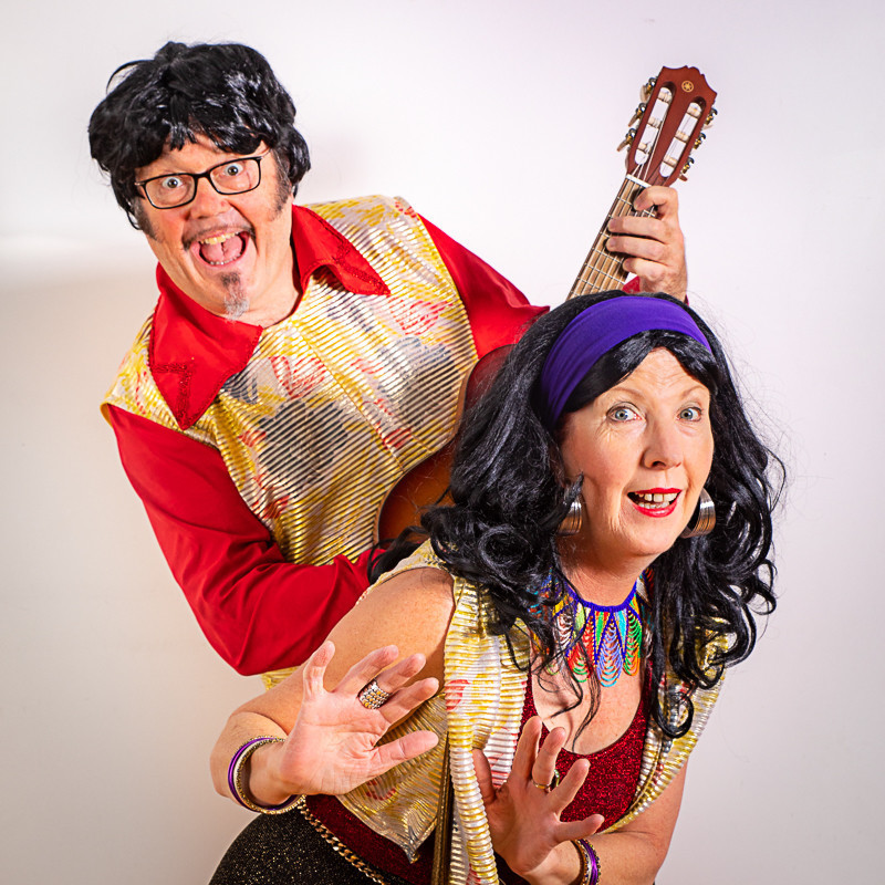 A photo of a man and a woman with amused facial expressions. The woman at the front has long black hair and is wearing a purple headband with a colourful necklace, red singlet top, and a yellow vest. The man behind her has short black hair and is wearing a red shirt, yellow vest and black framed glasses. He is holding a guitar.