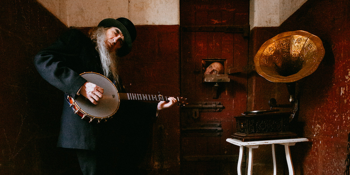 Outside the door of an old prison cell, an elderly man plucks a banjo. Opposite him is a  shiny brass gramophone. In-between these, visible through a tiny opening in the prison door, is the face of a second man, who looks out with consternation.