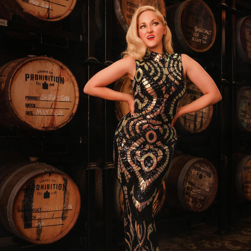Bootlegger Burlesque - Blonde woman in black and gold 1920s gown standing in front of gin barrels with 'prohibition' written across them.