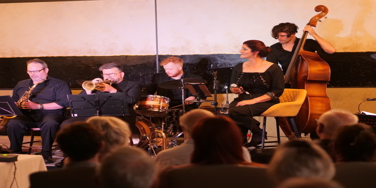 5 piece jazz band playing music, vocalist seated, singing wearing a black sparkly dress.