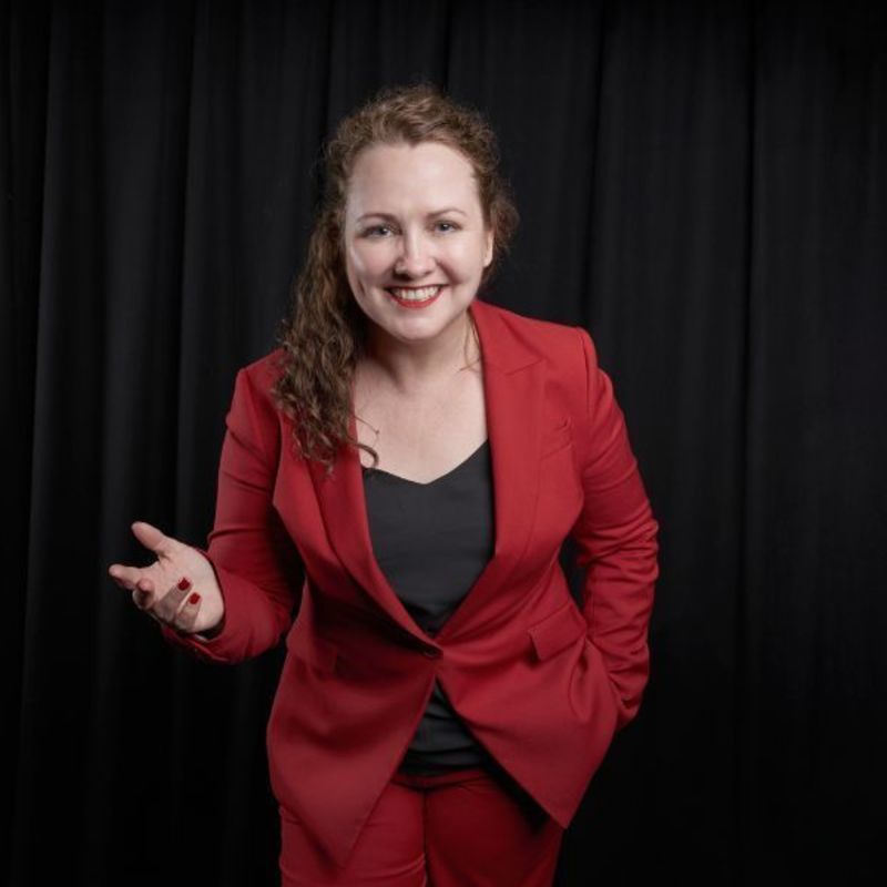 KC is a friendly-looking white woman in a red business suit, with a simple black top work under the jacket. She has curly brunette hair cascading over one shoulder and wears red lipstick. She is standing in front of a heavy black curtain and is lit as if by a spotlight.
