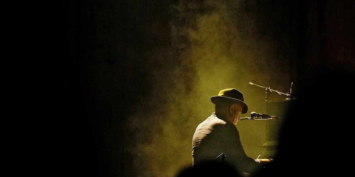 Greenish grey, smokey hues envelop Stewart D'Arrietta as he sits, with his back hunched, at his baby grand piano. He's facing right and is  turned away from us. The tone is dark and moody. We can't see his face, but we know it's him because he's wearing his signature felt hat; which is a type of fedora.