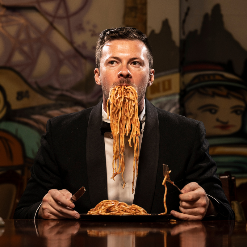 An image of Ben in a tuxedo, he is sitting at a dinner table alone with a knife and fork in hand. He appears to look quite formal, however there is an incredibly amount of spaghetti shoved into his mouth.