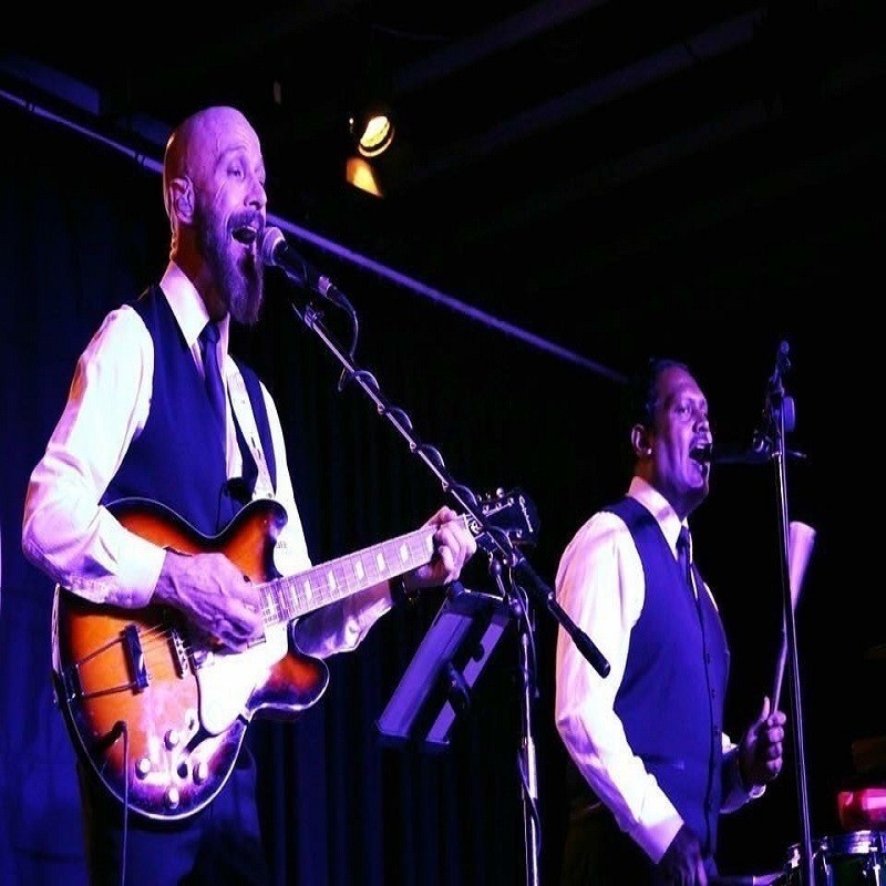 A photo of two men performing on stage singing into microphones. The man on the left is playing guitar.