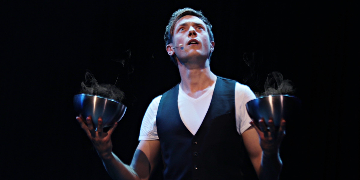 Magician Martin Brock holding two silver bowls with smoke coming out of them while looking curiously upwards.