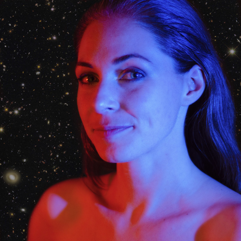 A headshot photo of a woman that has long brown hair slicked back. The background resembles the night sky with a black sky soft yellow stars.