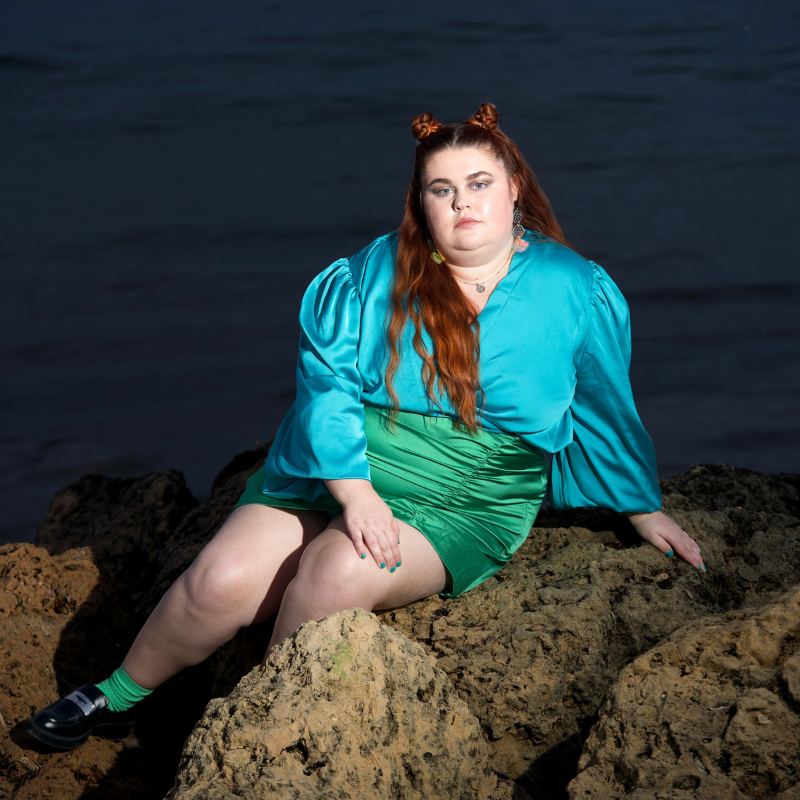 Image of Courtney [lead character and actor] sitting on a rock at the beach in a blue and green silk dress.