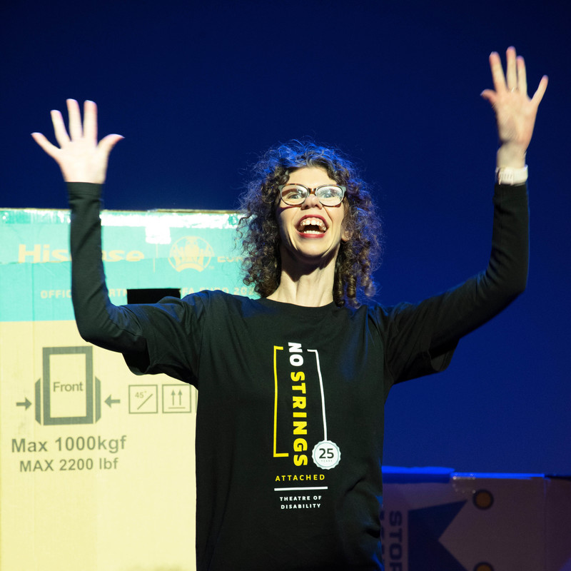 A woman with curly hair and glasses is on stage. She has raised both her arms out and her palms are facing towards you.