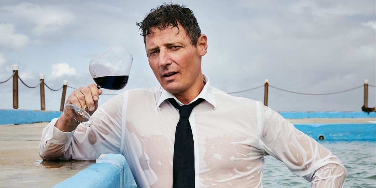 Merrick Watts - An Idiot's Guide to Wine - VOLUME TWO - Merrick stands in an outdoor pool, in a soaking wet white shirt, black tie holding a glass of red wine.