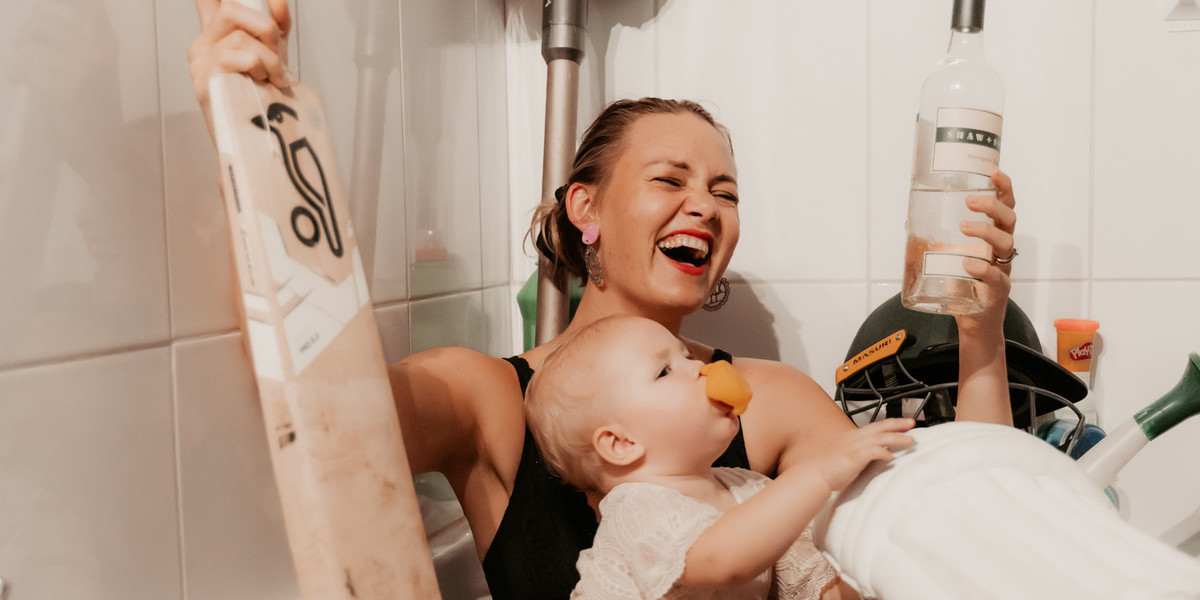 Women in a chaotic bath. there is a baby, cricket gear, juggling balls, vacuum cleaner and household products strewn everywhere.