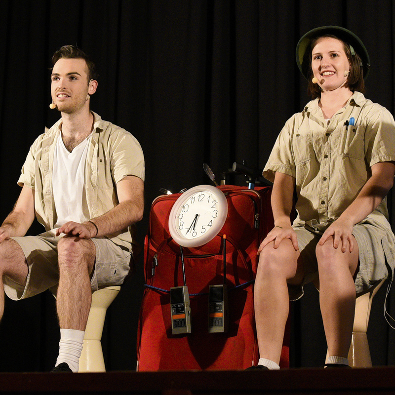 Two performers dressed as explorers sit on stools either side of a suitcase covered in toys and household objects dressed up to look like a machine