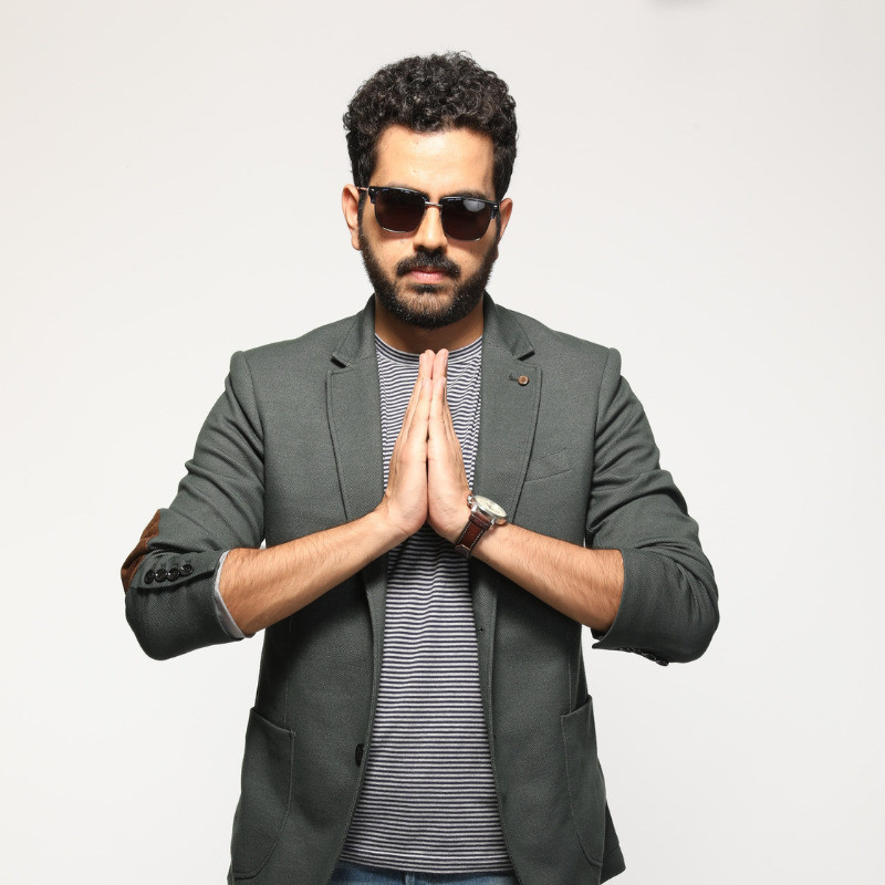 Performer wearing an open jacket (blazer), sunglasses, posing with the 'Namaste' sign
