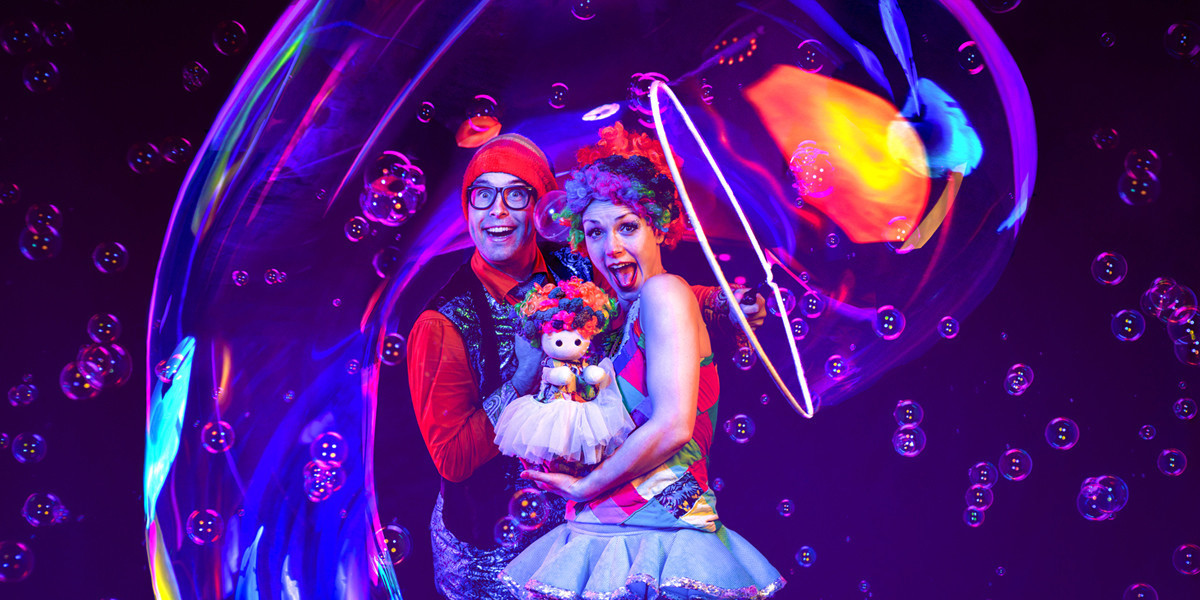 Three characters, a colourful doctor, a tiny rainbow haired puppet and a female clown stand together with big smiles across their faces whilst the doctor makes a long tube bubble that surrounds the three of them.