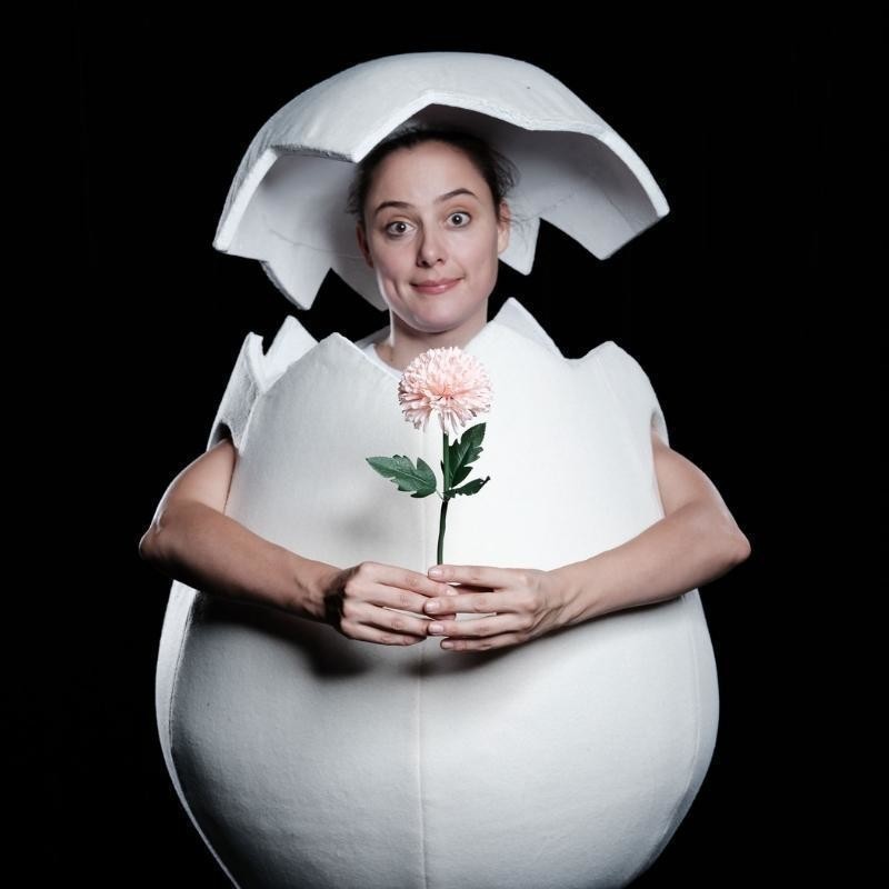 A single woman stands in a white round egg costume. She has white tights on and a white egg shell hat. She is holding a pink flower in her hands and has a sweet, closed lips smile on her face. She is staring straight to camera.