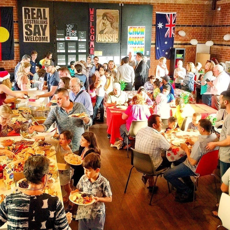 A photo of lots of people in a room standing and sitting. There is a long buffet table on one side with plates of food. There are multiple posters on the wall that say ‘Real Australians Say Welcome’ and ‘Aussie.