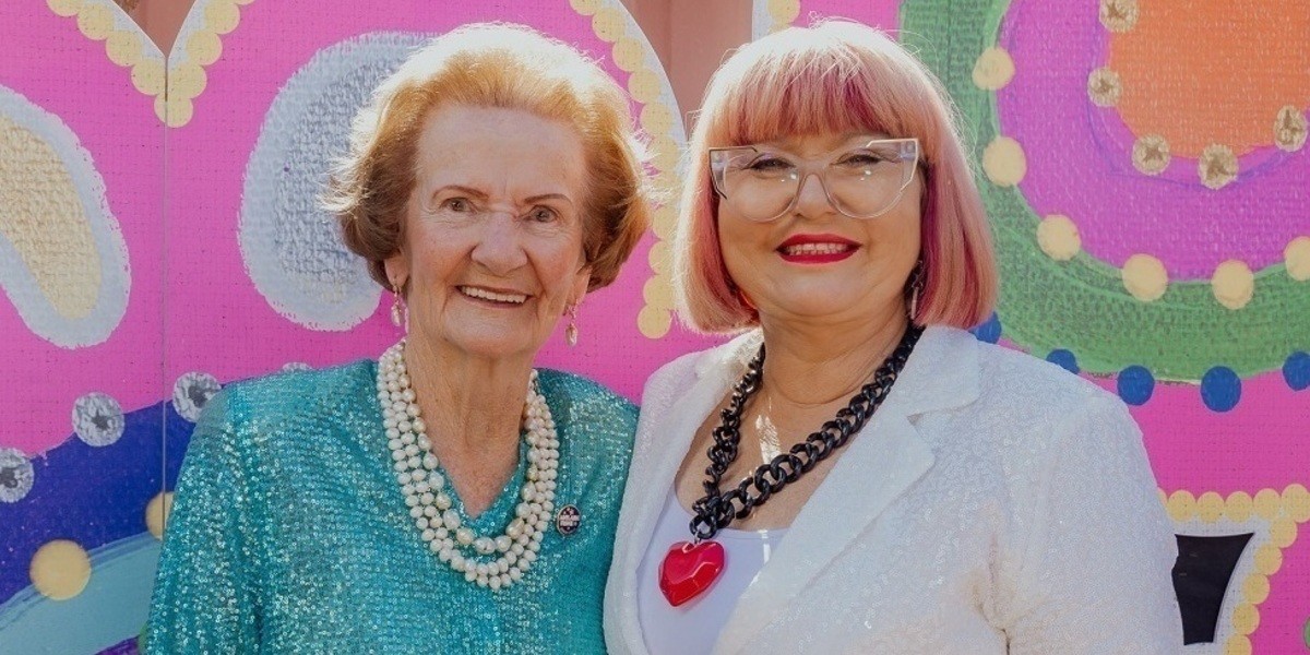 Sew Sparkly | The Gowns of The Mother of Fringe - MarjorieFitz-Gerald and Heather Croall are smiling directly at the camera. Marjorie wears a blue sequin bomber jacket and pearl necklace, Heather wears a white suit jacket and top with a red heart necklace.