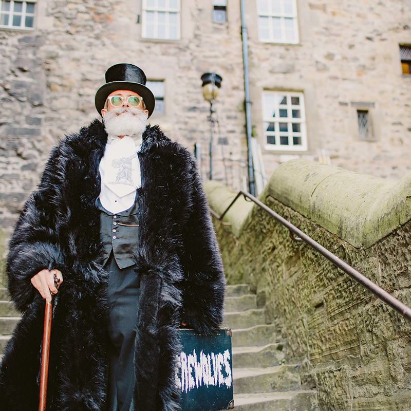 A bearded gentleman is descending a staircase outside of some old buildings in Edinburgh, he is wearing a top hat and a large black fur coat and is carrying a walking cane and a case with Werewolves written on it.