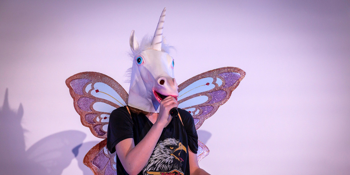 A person wearing a realistic unicorn mask and fairy wings, speaks into a microphone