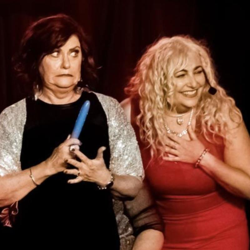 A woman on stage in a black dress holding a vibrator with an embarrassed impression. A woman in a red dress is laughing next to her.