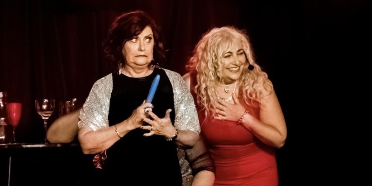 A woman on stage in a black dress holding a vibrator with an embarrassed impression. A woman in a red dress is laughing next to her.