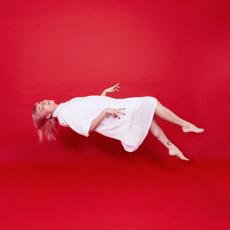 A worried looking woman with pink hair wears a white nightdress and levitates horizontally in front of a red background.
