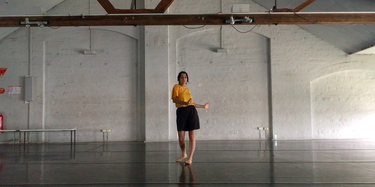 A lone dancer in a yellow t shirt and shorts strikes a pose in front of a blank white wall