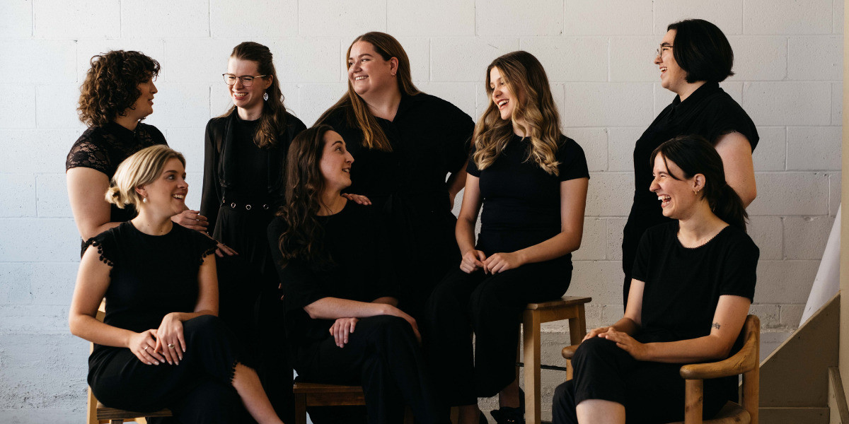 Vernacular - A group of women and non-binary people are sitting and standing around each other, talking. They are all wearing black.