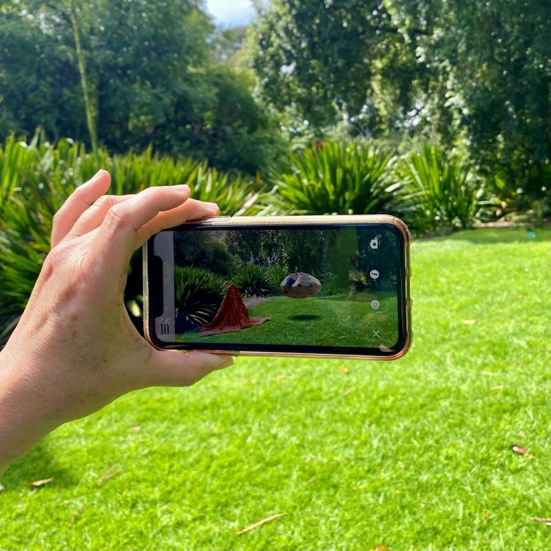 Seeing the Invisible at Adelaide Botanic Garden showing Augmented Reality artwork through smartphone of boulder suspended in the air and cloak made from bottle tops