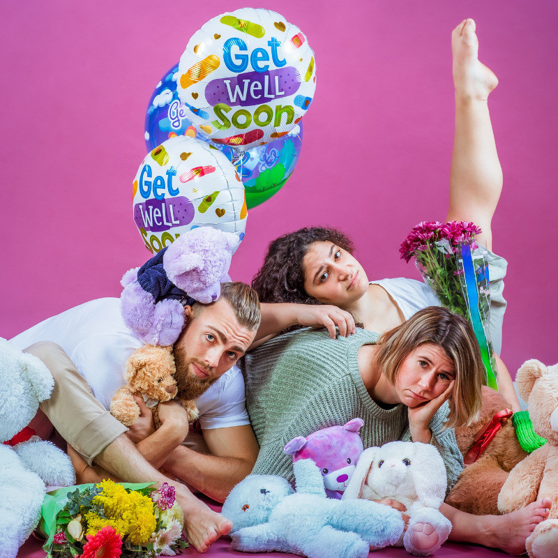 Three acrobats sit contorted and crumpled over each other on the floor with woeful facial expressions, set against a bright purple background. They are covered in teddy bears and a bouquet of flowers. Maxx on the left holds a ribbon tied to floating silver helium balloons which read "Get Well Soon". Emily on the far right has her leg up at a straight angle.