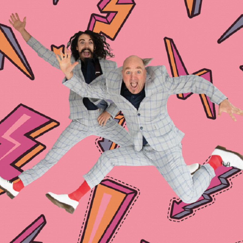 Two men wearing matching grey suits and jumping high in the air, against a pink background with colourful lightning bolts.