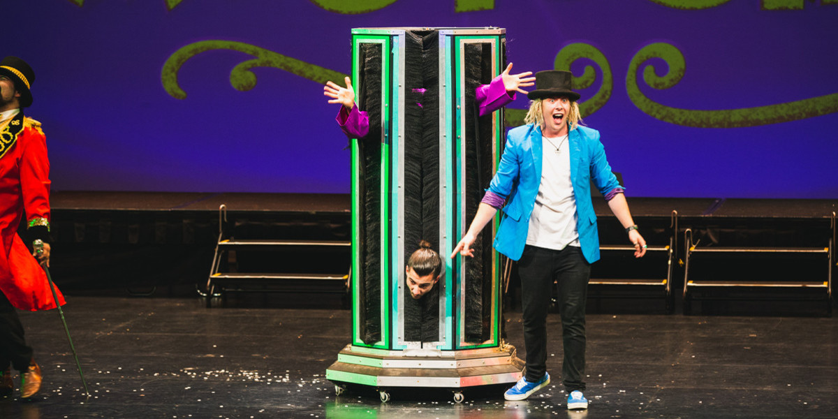 male performer inside a tall bow, head near the floor and two arms at head height. Another male performer pointing at the head and laughing.