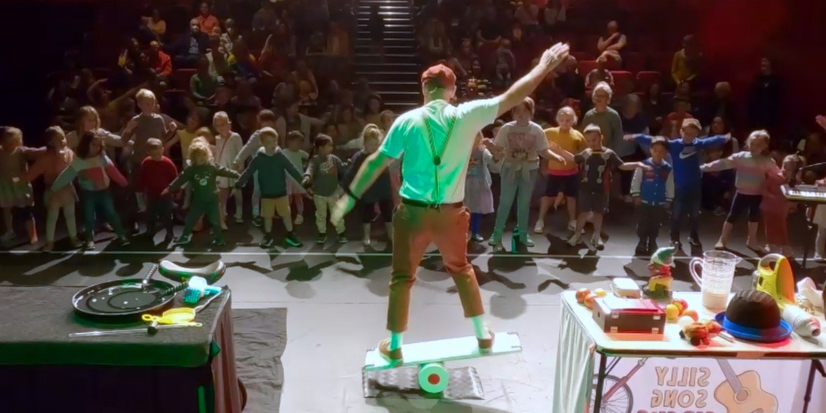 The artists is balancing on a board on top of a tube. He stands in front of a live audience with his back to the camera