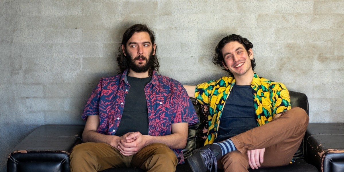 Two men sit on a couch wearing colourful shirts. The man to the left is looking off to the side and the man on the right is smiling.