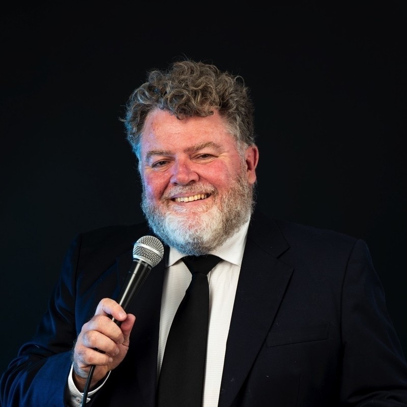 Coming Clean - A photo of a guy smiling at the camera holding a microphone to his house. He has short grey curly hair and a grey beard. He is wearing a black suit, black tie and white shirt.