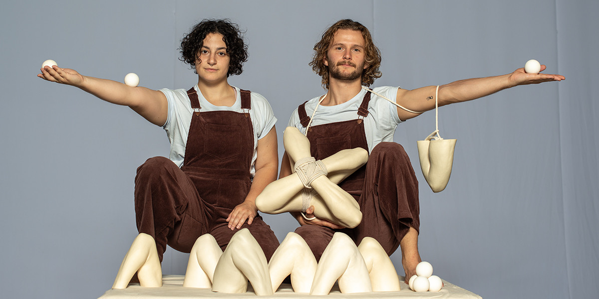 Both performers sit posed next to one another. They are surrounded by ceramic elbow sculptures. Emily holds two juggling balls one in their hand and one on their elbow, arm outstretched.