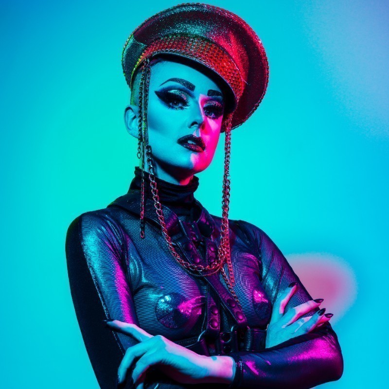 A photograph of a person wearing a long-sleeve black mesh top with a black harness over the top. They are also wearing an oversized gold glittery sailor cap. Their makeup has been heavily applied, featuring bold black eyeliner across their eyelids with gold eyeshadow and red glittery lipstick.