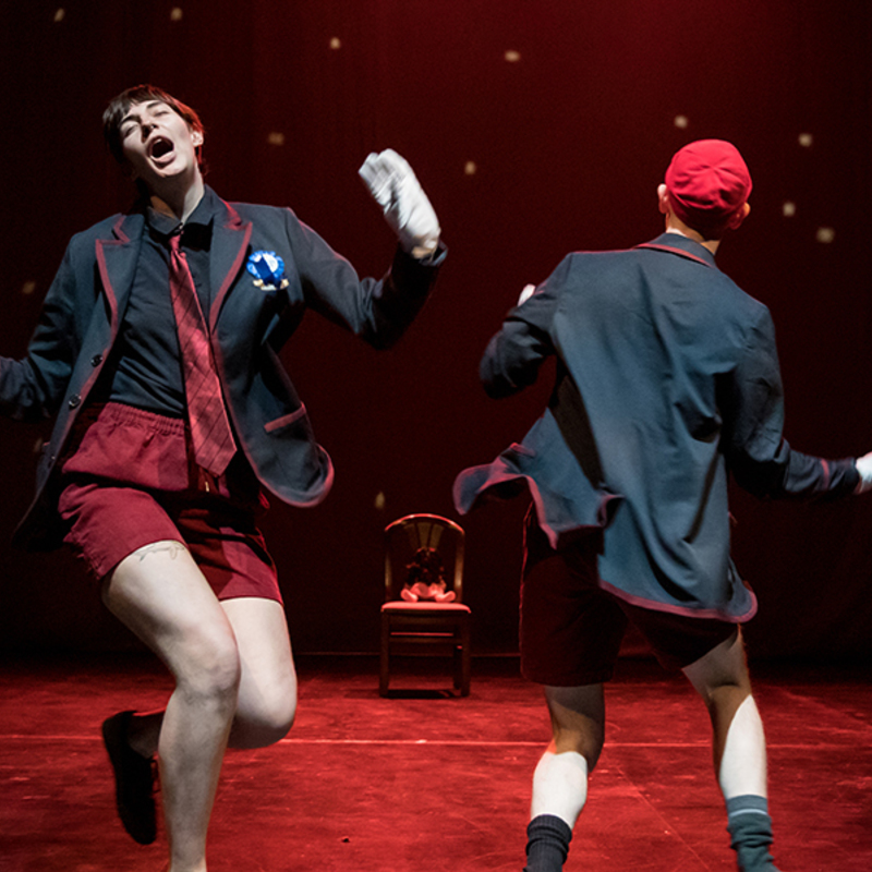 The two creepy boys are dancing under red light, waving their hands.  Both boys are dressed in dark blue blazers (similar to a child's school uniform) and maroon corduroy shorts.  They both are wearing small red touques, and white gloves.  The boy on the left (Grumms) is facing the camera, and the boy on the right (Sam) is facing away from the camera.  In the background there is a wooden chair.  The stage is lit with red light.