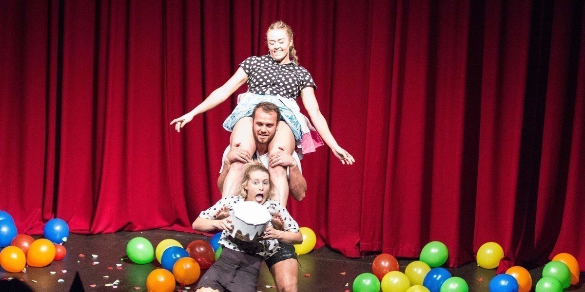 Three performers in casual clothes, stacked on top of each other, balance a cake in front of a red curtain and a floor covered in multicolored balloons.