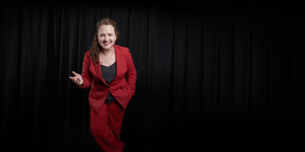 Archaeologist / comedian, KC Martin-Stone, stands in front of black theatrical curtains, with a friendly smile on her face and an open gesture with her right hand. She is wearing a striking red pants suit and a simple black top.