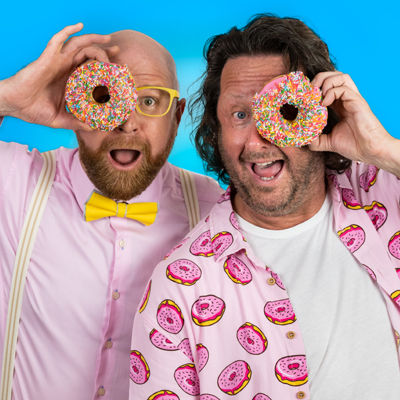 A bald man in a pink shirt, braces and yellow bow tie looks through the centre of a doughnut. Next to him, a man in a white t-shirt and pink shirt looks through the centre of a doughnut.
